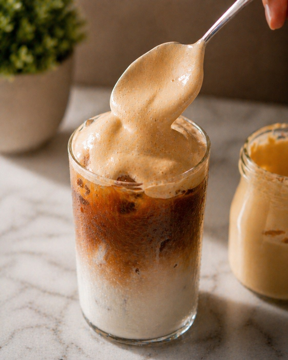Spooning thick caramel cold foam onto the layered iced coffee — the foam piled high on a spoon