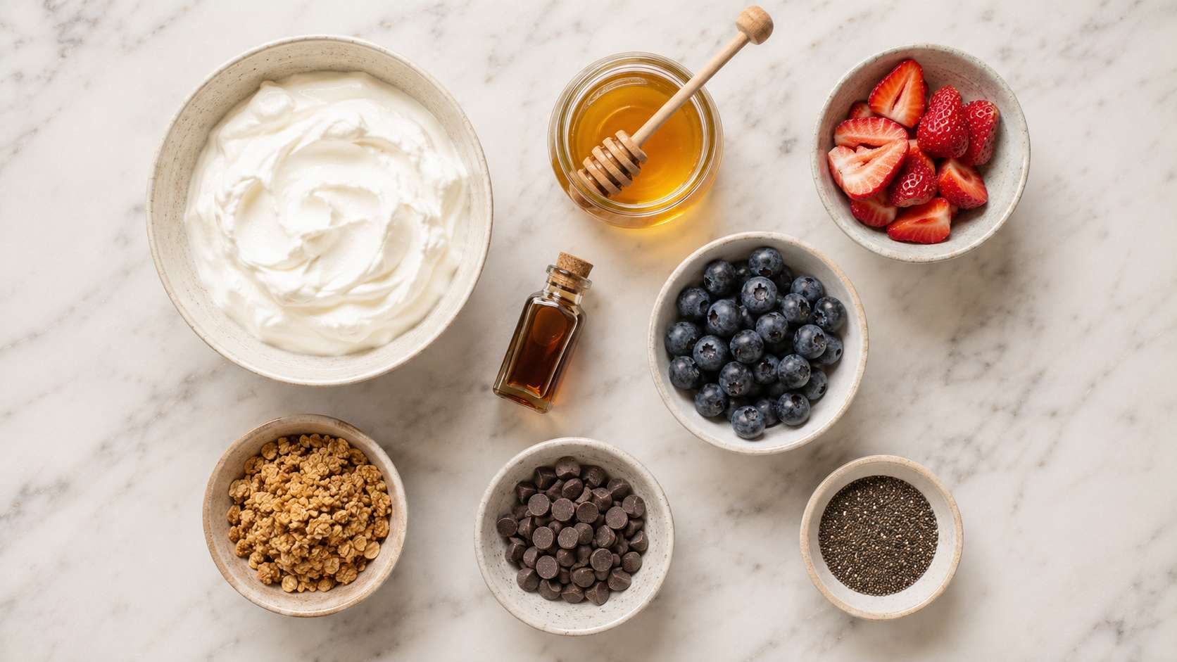 All yogurt bark ingredients laid out: Greek yogurt, honey, vanilla, strawberries, blueberries, granola and chocolate chips