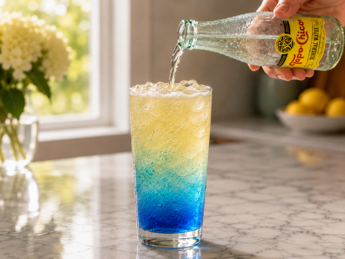 Sparkling water poured gently over the top of the blue layered mocktail — bubbles rising through the color