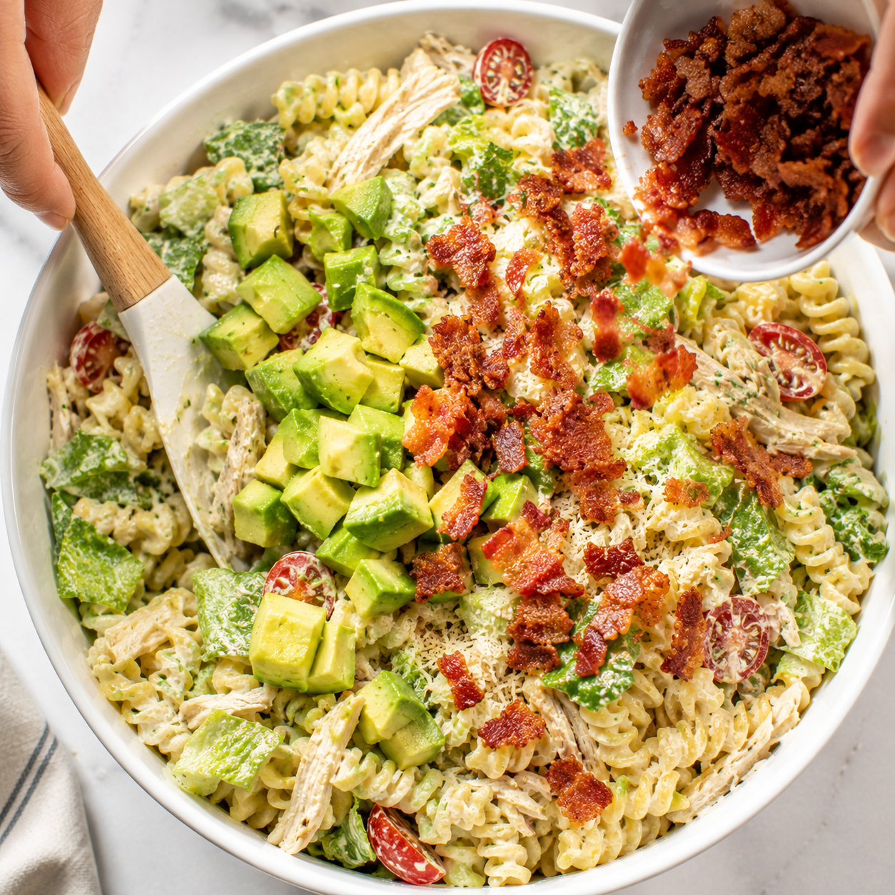 Diced avocado and crumbled bacon being gently folded into the pasta salad