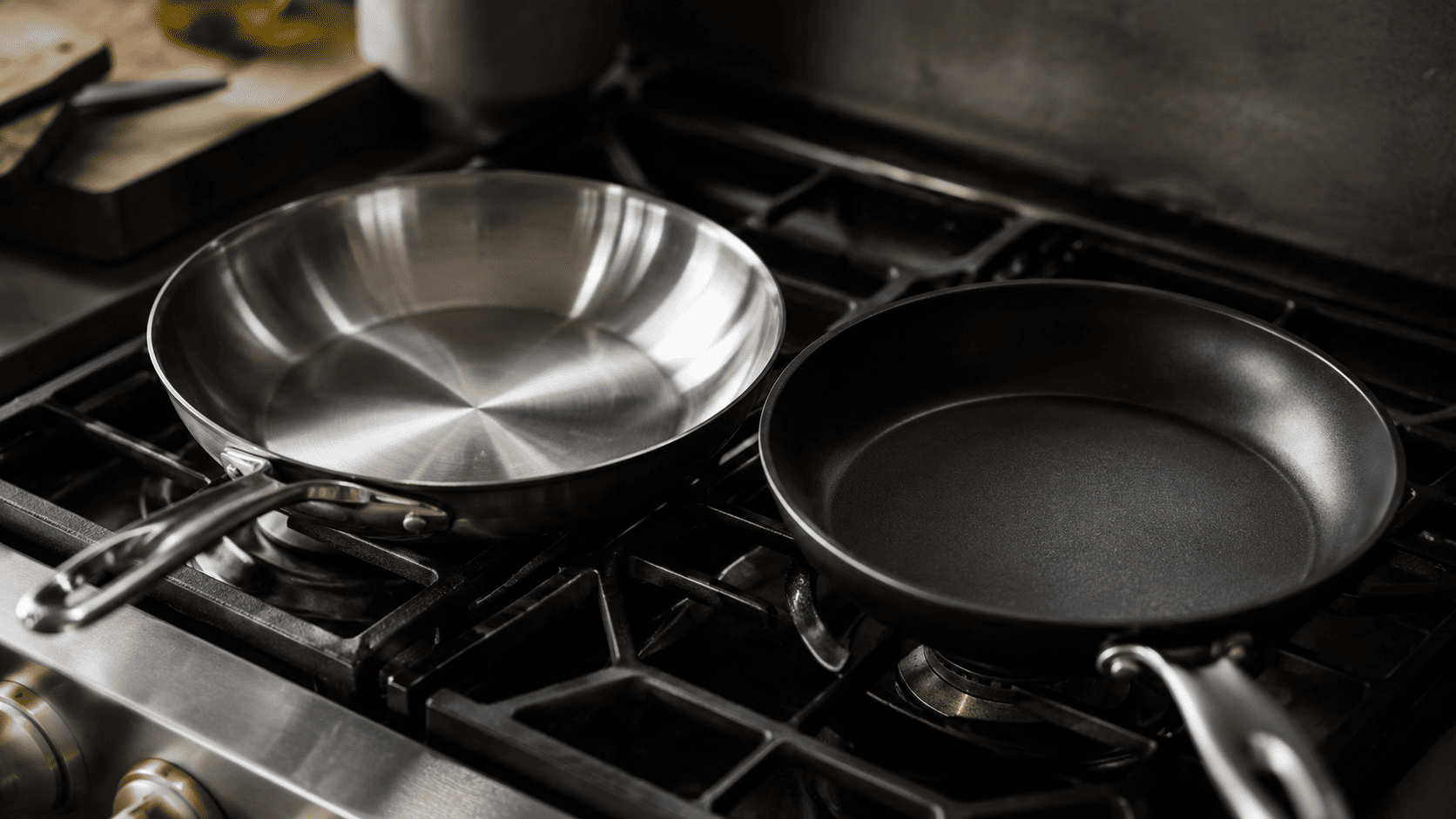 A brushed stainless steel skillet and a dark non-stick frying pan side by side on a stovetop, moody editorial lighting