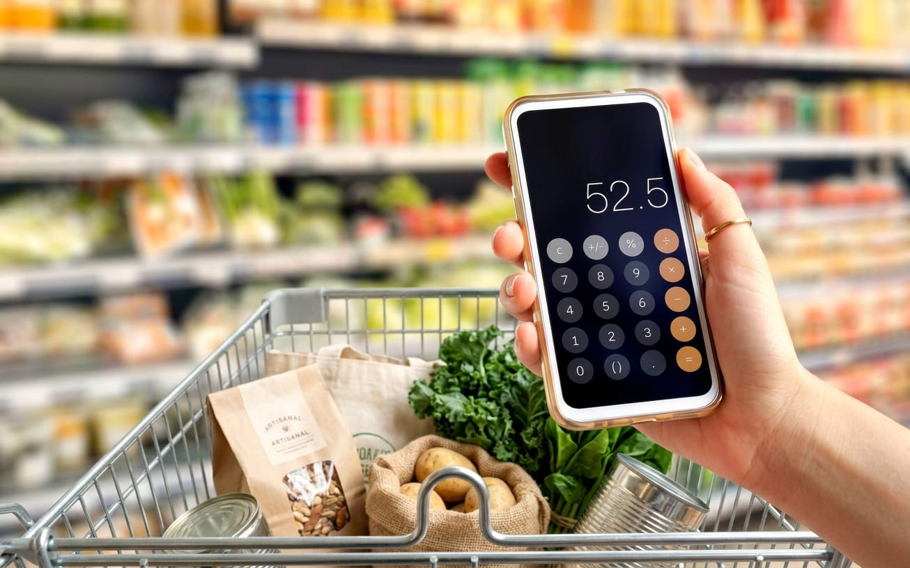 A hand holding a phone calculator app over a half-full grocery cart, supermarket aisle softly blurred behind, natural overhead light