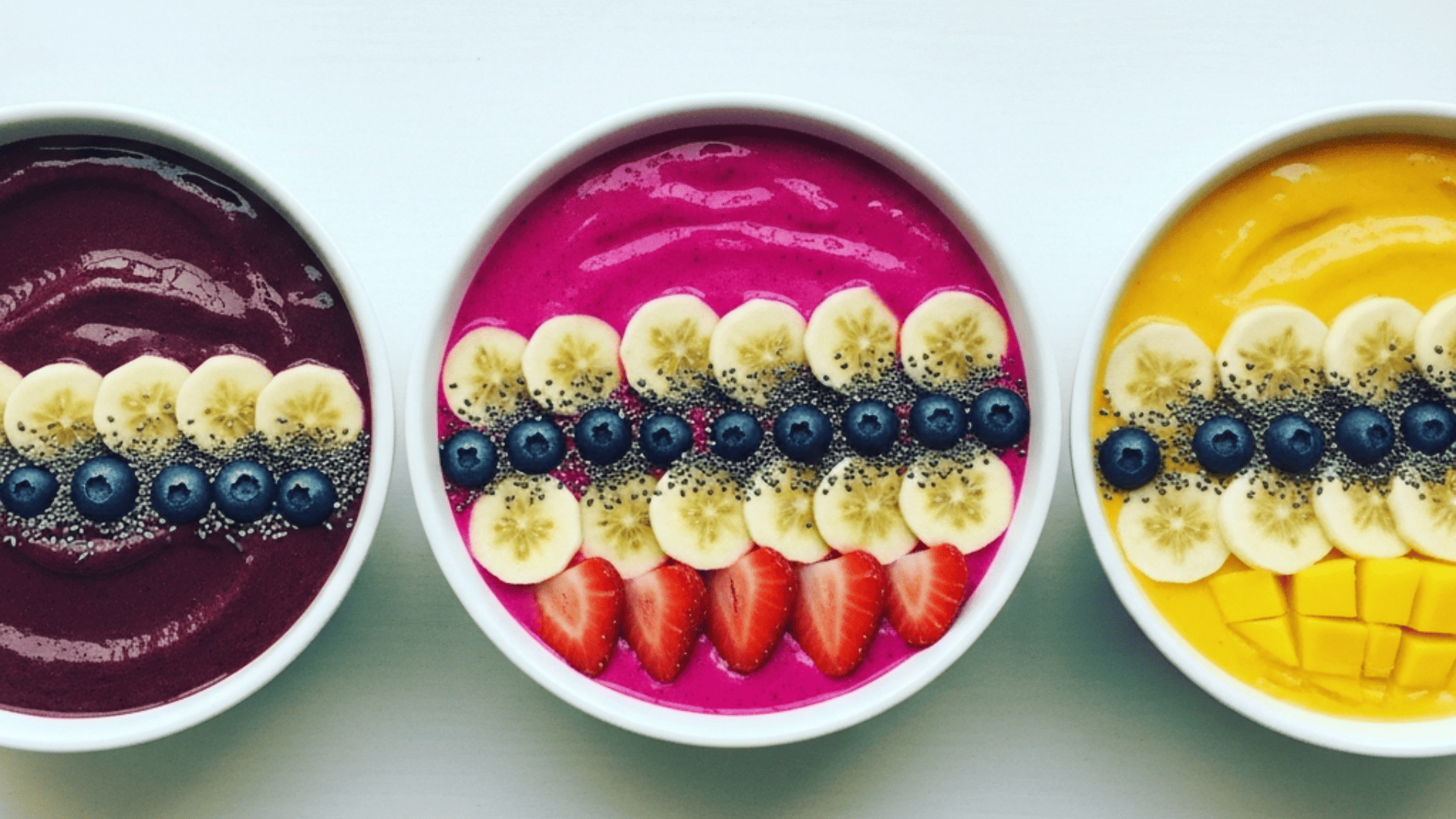 An overhead row of three vivid smoothie bowls — deep purple açaí, hot-pink pitaya, golden mango — on a pale ceramic surface, toppings arranged in neat rows, soft morning light