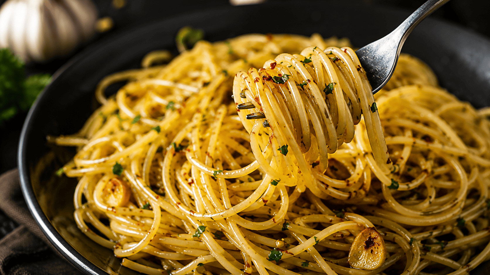 Glossy spaghetti coated in golden garlic oil, flecks of chili and parsley, twirled on a fork over a dark plate