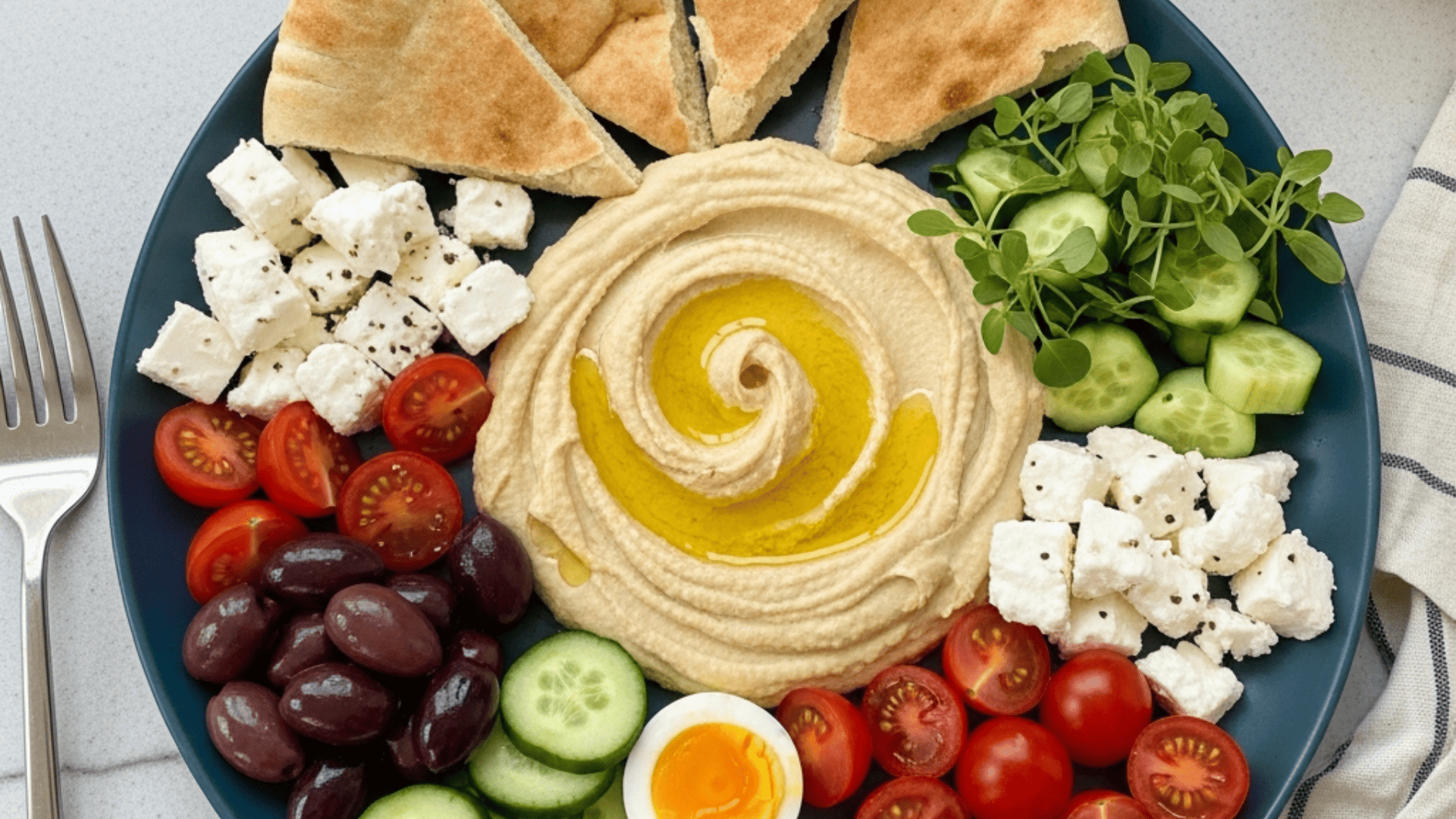 Mediterranean mezze brunch board — hummus with an olive-oil swirl, warm torn pita, olives, feta, cherry tomatoes, cucumber, halved soft-boiled eggs and fresh herbs