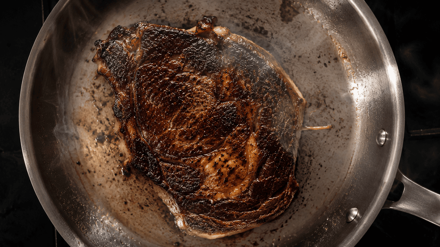 Overhead shot of a deeply seared steak crust in a stainless steel skillet, cast iron-dark sear marks, steam rising