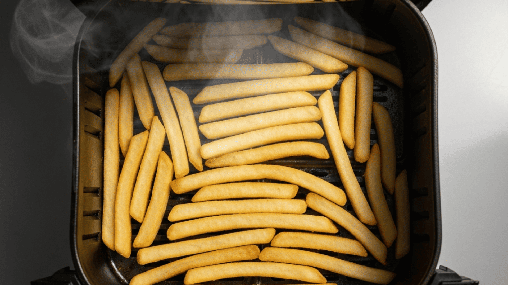 Overhead shot of golden, evenly crisped fries in a single layer inside an air fryer basket with warm directional light and steam
