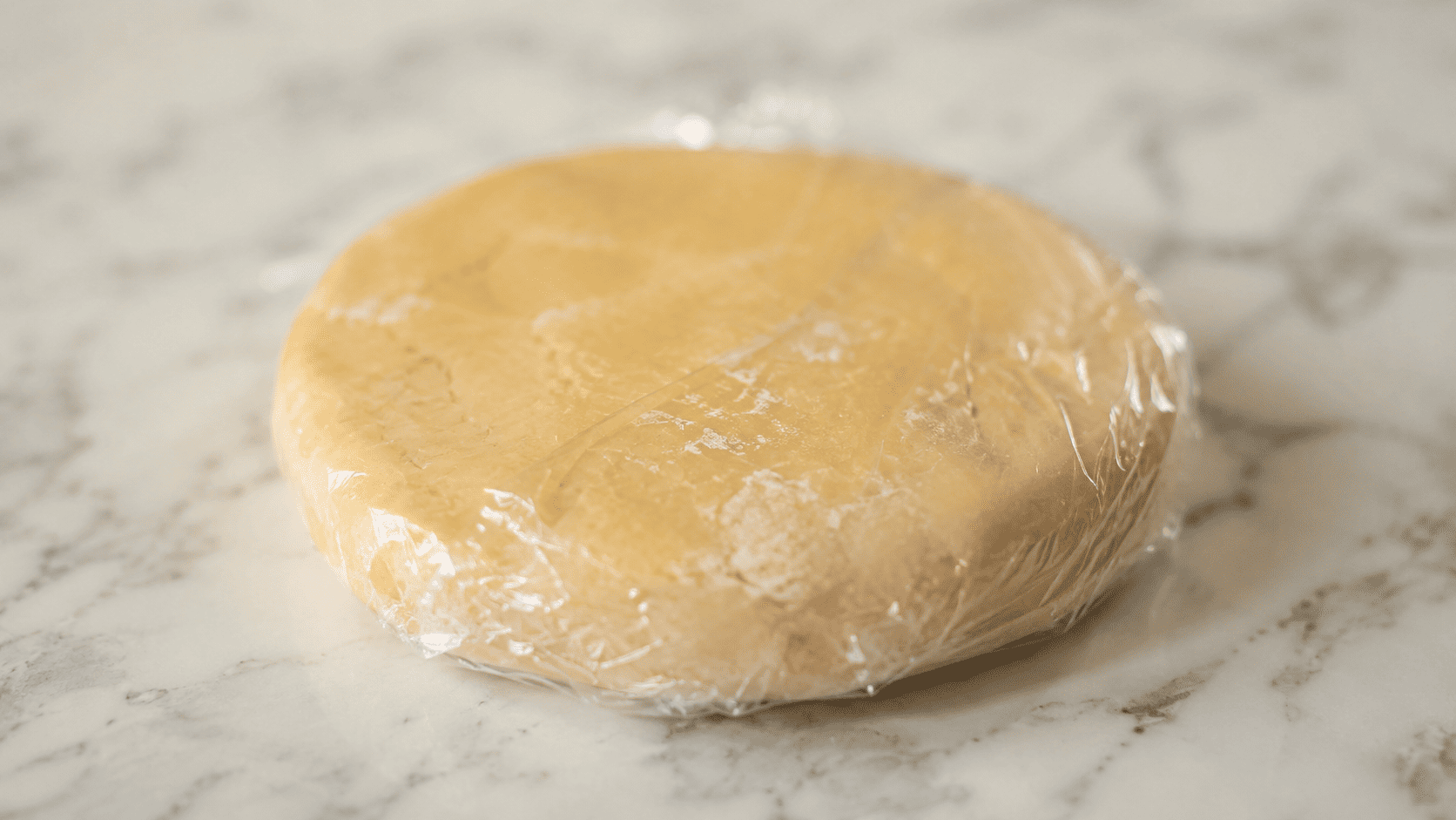 A disc of pastry dough wrapped in plastic wrap resting on a white marble counter, chilling before rolling