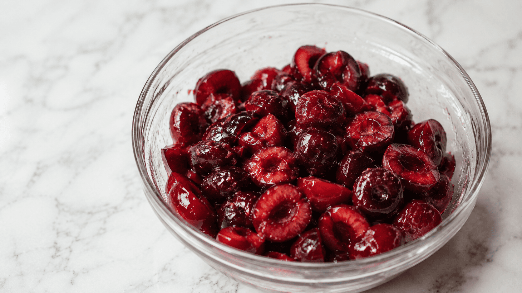 A glass bowl of fresh halved cherries glistening with released juice and sugar, ready for the galette filling