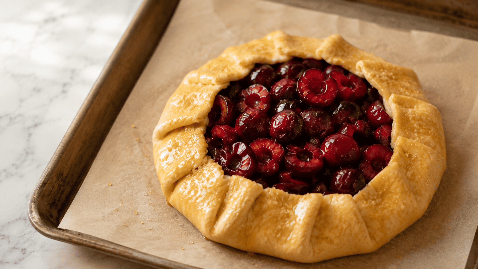 An assembled cherry galette on parchment paper on a baking sheet, crust brushed with glossy egg wash and sprinkled with coarse sugar, ready for the oven
