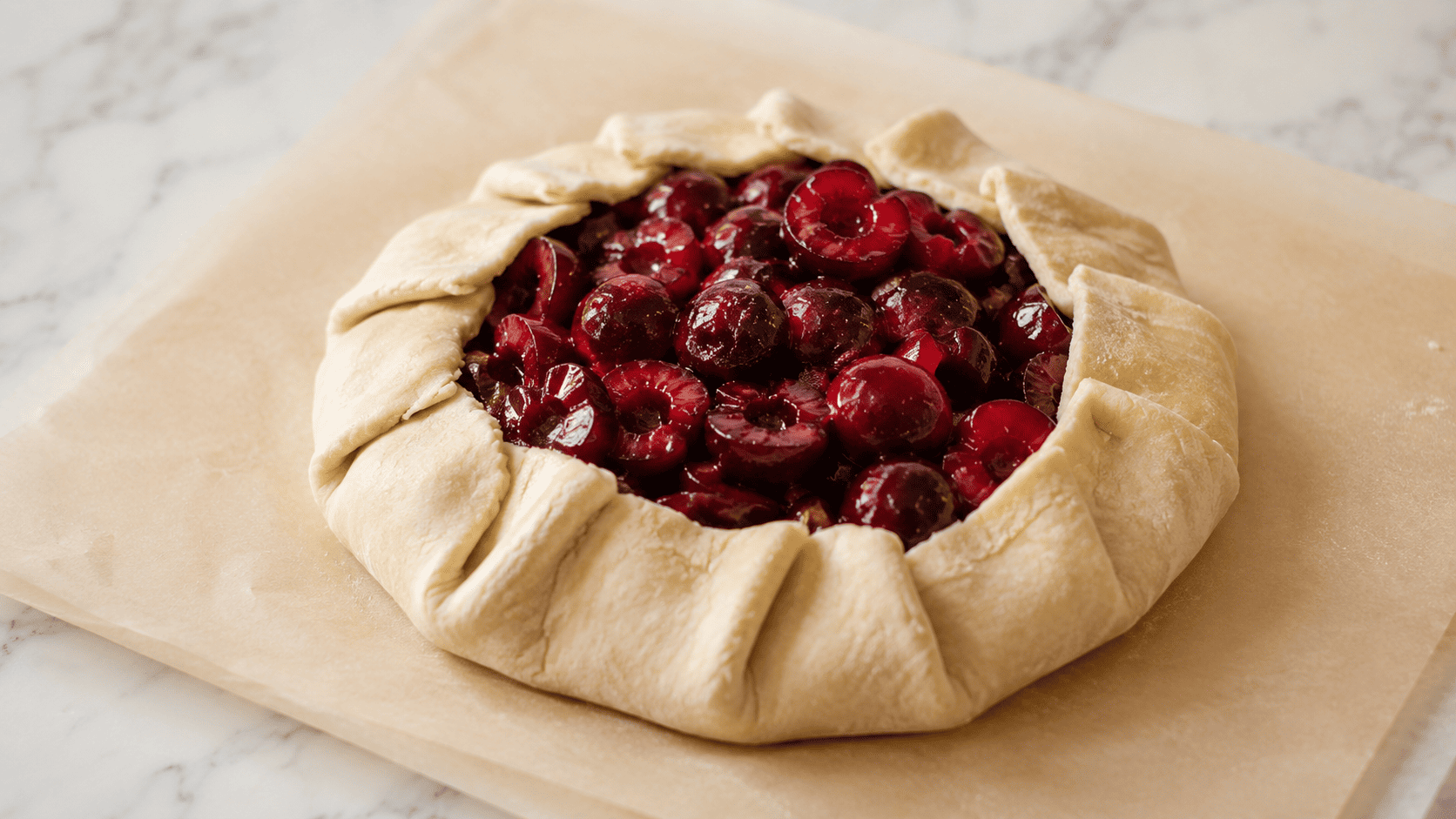 An unbaked galette on parchment paper on a white marble counter, cherry filling piled in the center before folding