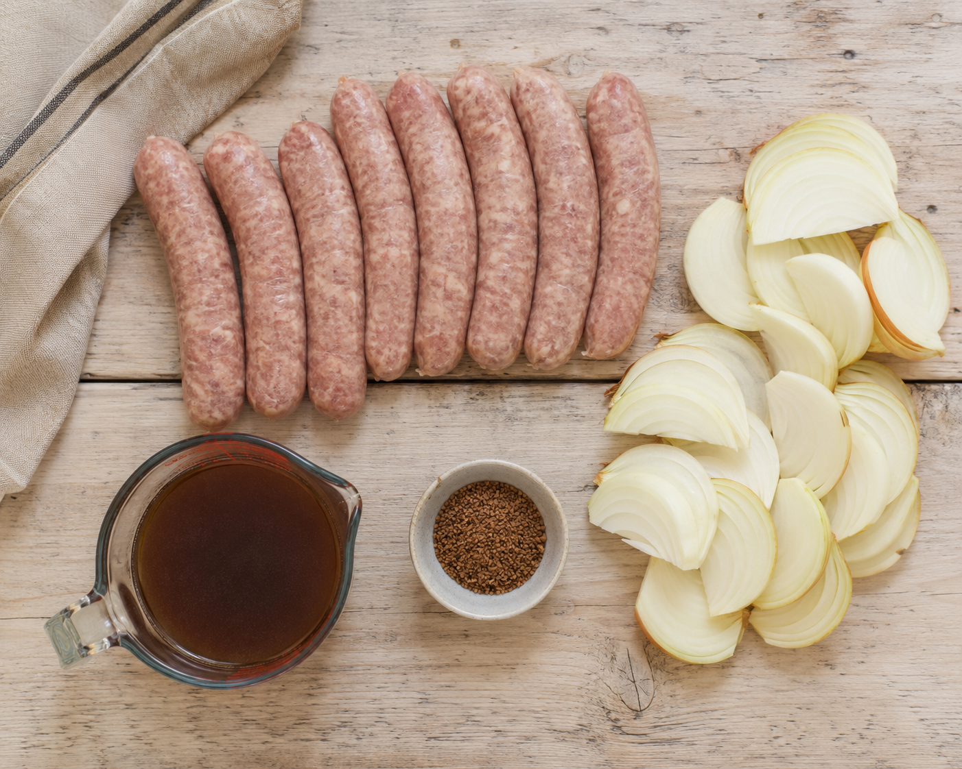 Flat-lay of raw pork sausages, sliced onions, beef stock, and gravy granules on a wooden board