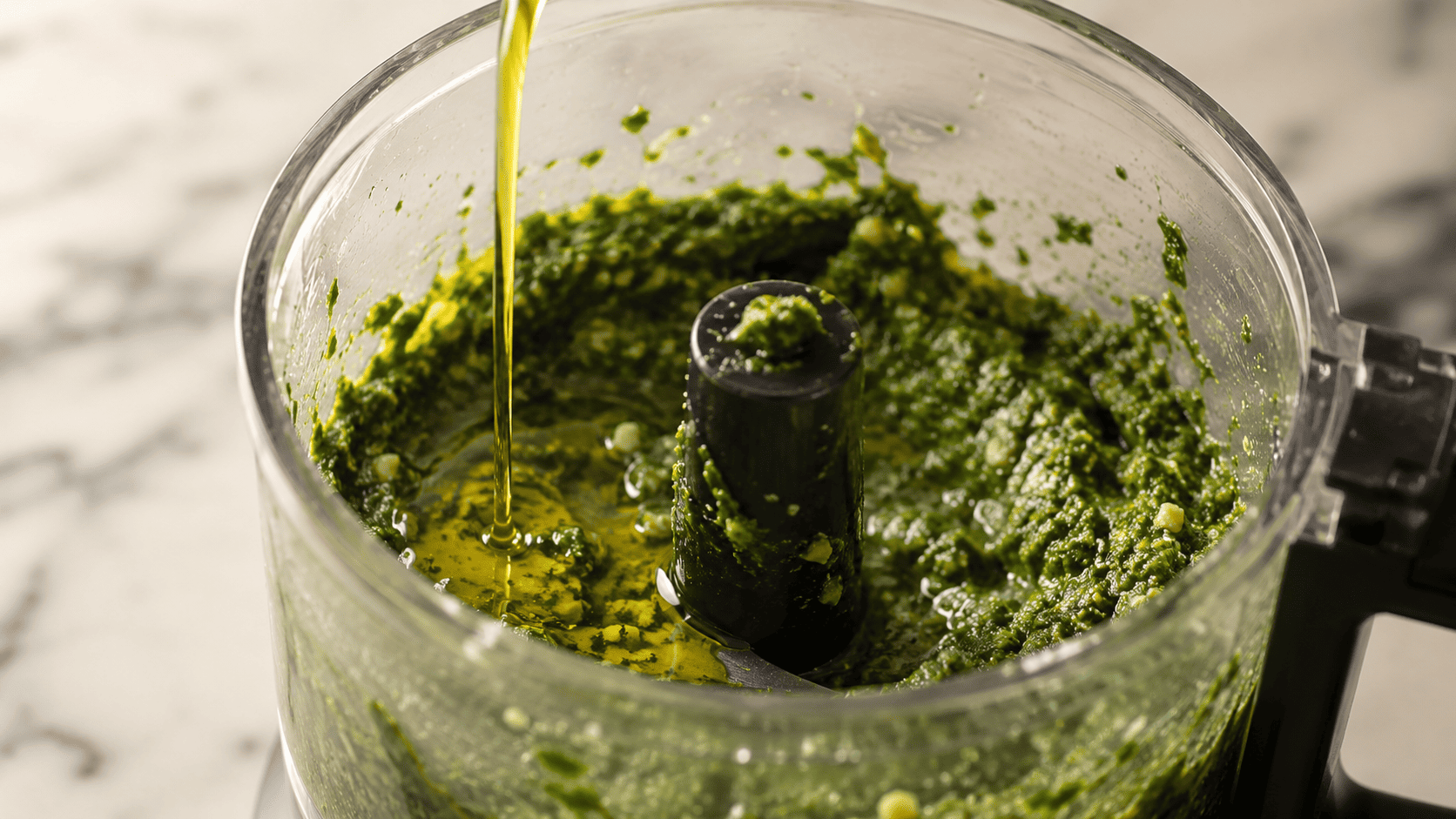 Olive oil being streamed into the food processor as the basil mixture turns into a bright green paste