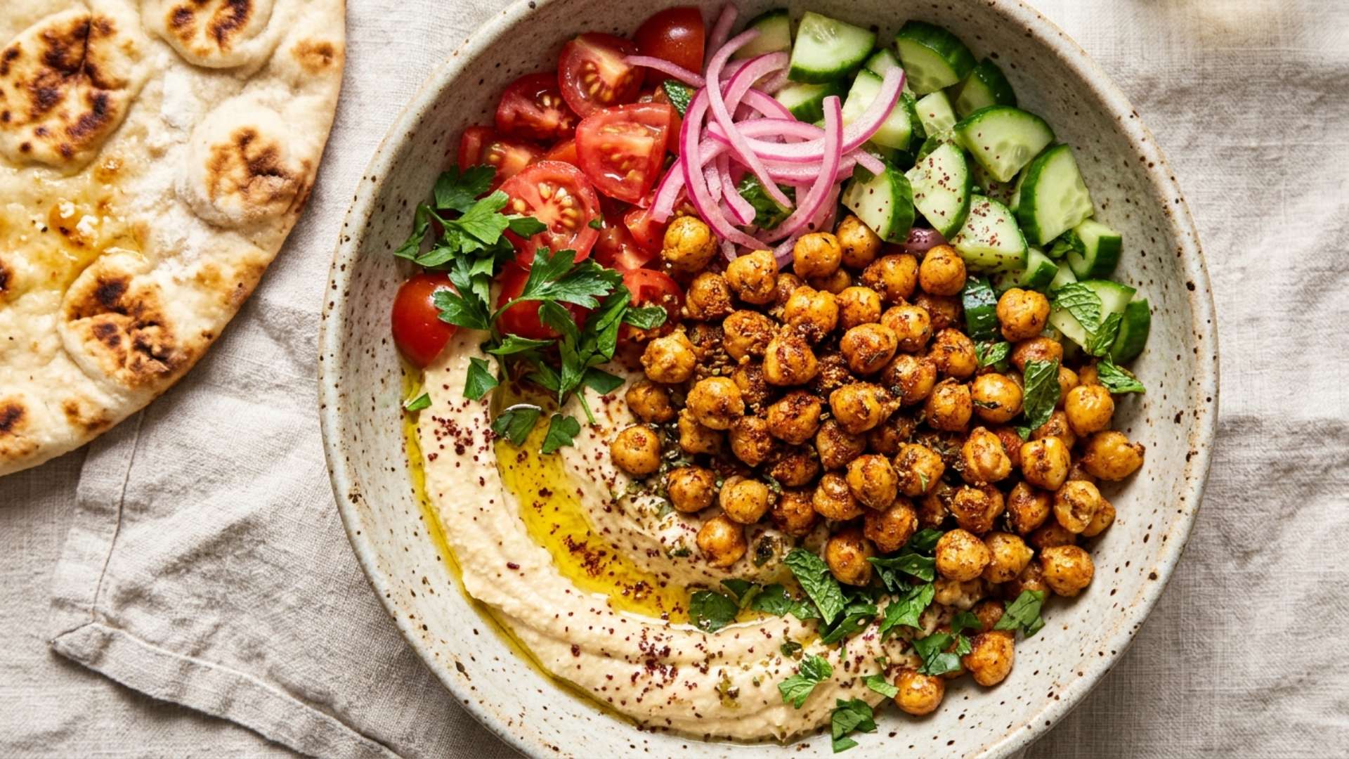 Bowl of crispy spiced chickpeas over hummus with cucumber, tomato and pickled onions