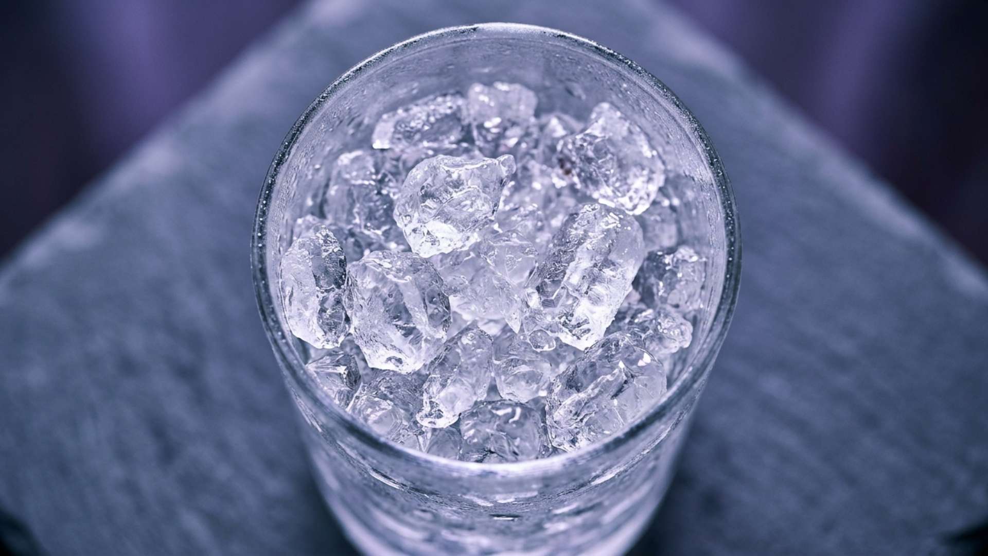 A glass tumbler full of crushed ice on a cool surface in violet light